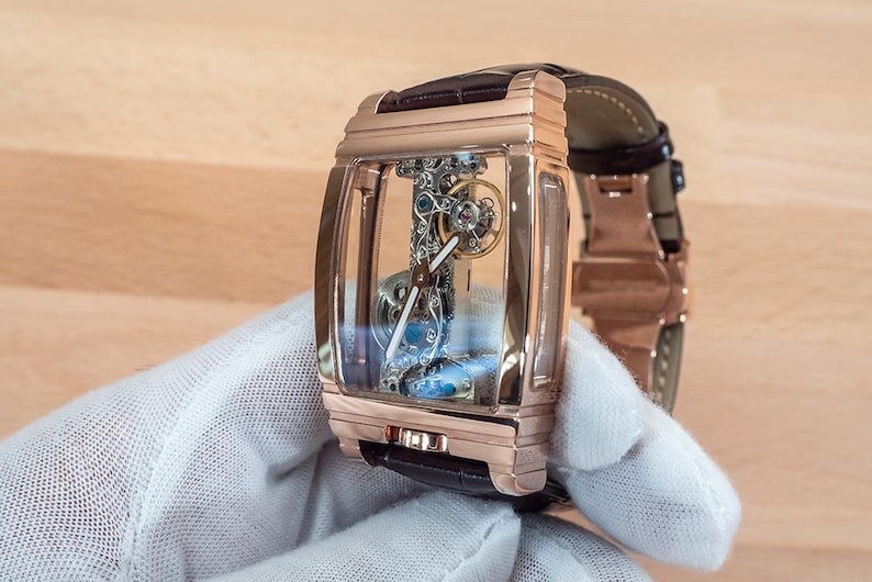 May include: A rose gold rectangular wristwatch with a transparent face, showcasing the intricate internal mechanisms. The watch has a brown leather strap and is held by a gloved hand. The watch's design is modern and luxurious.