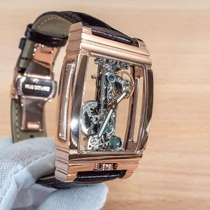 May include: A rose gold rectangular skeleton watch with a brown leather strap. The watch's intricate internal mechanisms are visible through the transparent face. The watch is held by a gloved hand.