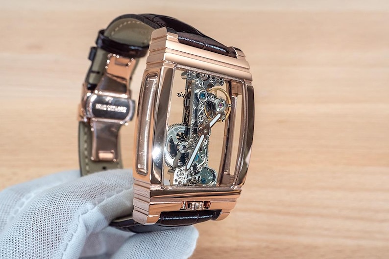 May include: A rose gold rectangular skeleton watch with a brown leather strap. The watch's intricate internal mechanisms are visible through the transparent face. The watch is held by a gloved hand.