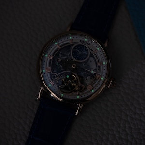 May include: A silver and rose gold skeleton watch with a blue leather strap. The watch face has a white background with black Roman numeral hour markers and a moon phase indicator. The watch is illuminated with green glow-in-the-dark accents.
