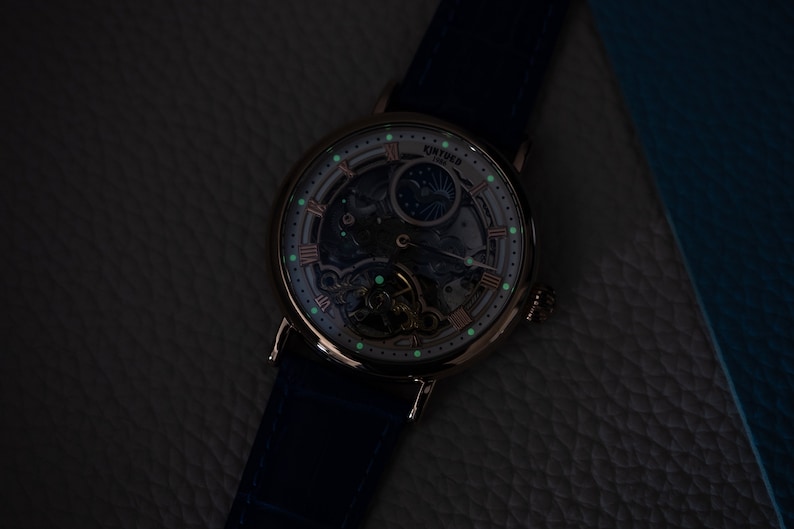 May include: A silver and rose gold skeleton watch with a blue leather strap. The watch face has a white background with black Roman numeral hour markers and a moon phase indicator. The watch is illuminated with green glow-in-the-dark accents.