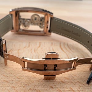May include: A rose gold wristwatch with a transparent face, revealing the intricate inner workings. The watch has a black leather strap and a rose gold clasp. The watch is displayed open on a wooden surface.
