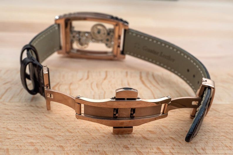 May include: A rose gold wristwatch with a transparent face, revealing the intricate inner workings. The watch has a black leather strap and a rose gold clasp. The watch is displayed open on a wooden surface.