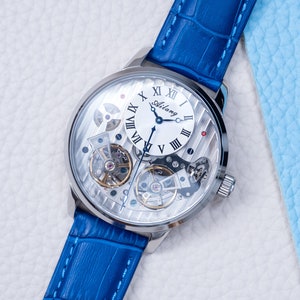 May include: A silver-toned wristwatch with a white face and Roman numeral hour markers. The watch has a blue leather band and a skeletonized design, revealing the inner workings of the watch.