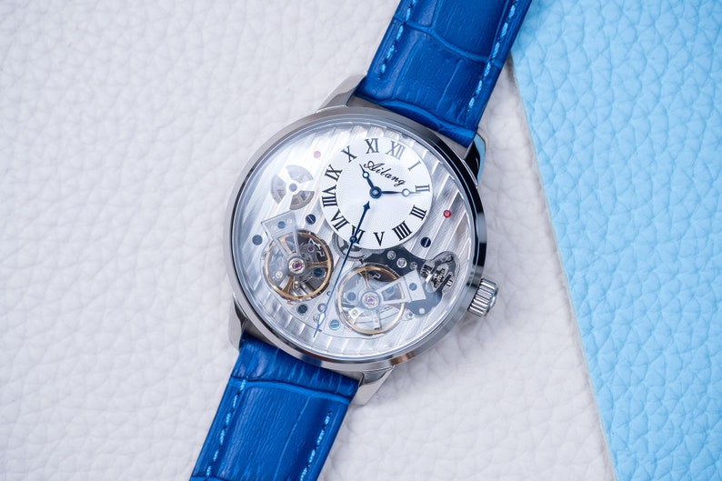 May include: A silver-toned wristwatch with a white face and Roman numeral hour markers. The watch has a blue leather band and a skeletonized design, revealing the inner workings of the watch.