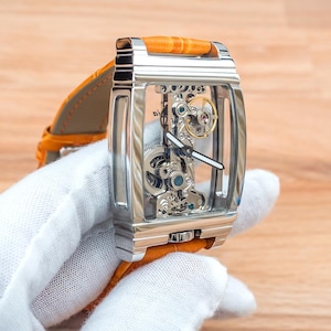 May include: A rectangular, silver-toned skeleton watch with a transparent face, showcasing the intricate inner workings. It features a bright orange leather band. The watch is held by a gloved hand, set against a wooden background.