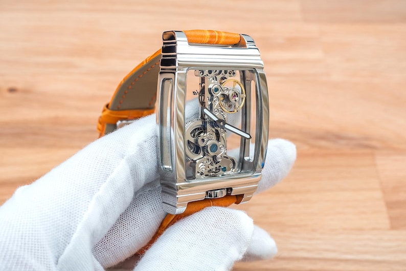 May include: A rectangular, silver-toned skeleton watch with a transparent face, showcasing the intricate inner workings. It features a bright orange leather band. The watch is held by a gloved hand, set against a wooden background.