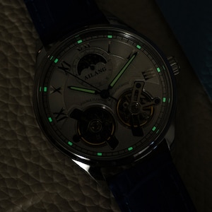 Pu&ograve; includere: Un orologio argentato con quadrante bianco e lancette nere. L'orologio ha un indicatore di fase lunare e un movimento scheletrato. L'orologio brilla di verde al buio.