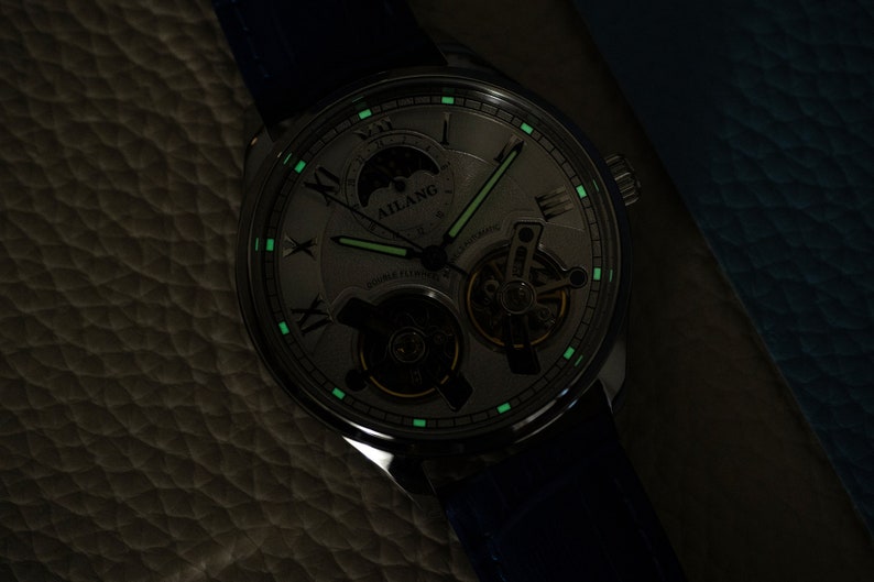 Pu&ograve; includere: Un orologio argentato con quadrante bianco e lancette nere. L'orologio ha un indicatore di fase lunare e un movimento scheletrato. L'orologio brilla di verde al buio.