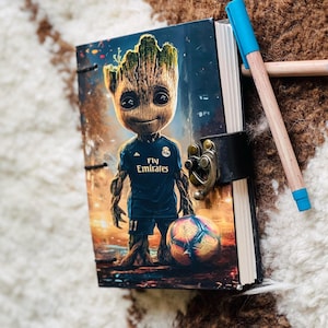May include: A black leather journal with a colorful image of a fictional character wearing a soccer jersey with the text "Fly Emirates". The journal has a metal clasp and a brown leather strap.