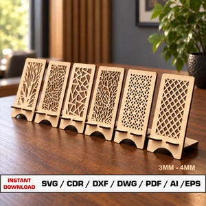 May include: Six light-colored wooden phone stands with intricate laser-cut geometric and organic patterns. Each stand features a small horizontal ledge for device support. Text at the bottom indicates 3MM - 4MM thickness and file formats.