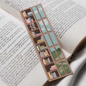 Puede incluir: Un marcapáginas marrón con un diseño de punto de cruz de una estantería llena de libros coloridos. La estantería está situada sobre un fondo de cielo azul y árboles verdes. El marcapáginas también incluye el texto "2023".