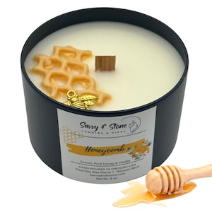 May include: A black candle jar with a white wax candle inside. The candle is decorated with a gold honeycomb design and a gold bee charm. The jar has a wooden lid and a label that reads "Savvy & Stone Candles & Gifts Honeycomb Scents: Pure Honey & Vanilla Hand Poured in Oregon | USA Para-Soy Wax Blend | Wooden Wick SavvyandStone.com Net Wt. 6 oz."