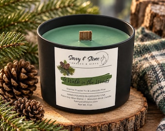May include: A matte black candle jar with green wax and a wooden wick. The white label features the text Savvy & Stone Candles & Gifts, A Walk in the Woods, Scents: Frasier Fir & Lakeside Pine, Hand Poured in Oregon, USA, and Net Wt. 6 oz.