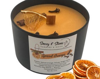 May include: A black candle jar with a gold lid and a label that reads "Savvy & Stone Candles & Gifts Spiced Orange". The candle is scented with spiced orange, cloves, and ginger spice. It is hand-poured in Oregon, USA, and made with a para-soy wax blend and a wooden wick. The net weight is 6 oz.