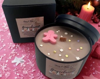 May include: A black tin candle with a gingerbread scent, featuring a pink gingerbread man and decorative sprinkles. The candle is in a black box with the text "Gingerbread" on the label. A lit candle and Christmas tree are in the background.