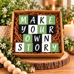 May include: A square cardboard gift box filled with brown paper crinkle shred containing sixteen wooden blocks arranged in four rows. The blocks feature white, black, and sage green surfaces with white or black lettering spelling out MAKE YOUR OWN STORY.