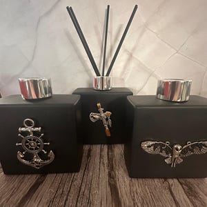 May include: Three black glass diffuser bottles with silver lids and decorative metal accents. The bottles are arranged in a row on a wooden surface. The accents include an anchor with a wheel, a Native American tomahawk with feathers, and an owl with outstretched wings.