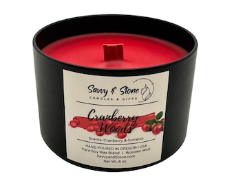 May include: A black candle container with a vibrant red wax interior and a wooden wick. The label reads "Cranberry Woods" with scents of cranberry and currants. The candle is hand-poured in Oregon, USA, and weighs 6 oz.