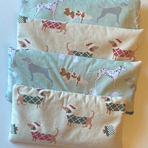 May include: Four different fabric-covered heat packs with a Christmas dog theme. The heat packs are stacked on top of each other. The top heat pack has a light green background with a white and brown dog wearing a red Santa hat. The second heat pack has a light beige background with a brown and white dog wearing a red Santa hat. The third heat pack has a light green background with a white and black dog wearing a red Santa hat. The bottom heat pack has a light beige background with a brown and white dog wearing a green Santa hat.