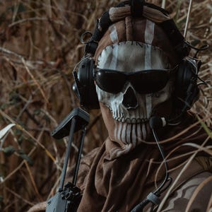 Puede incluir: Una persona con equipo táctico, que incluye una máscara con forma de calavera, gafas de sol y auriculares, sosteniendo un rifle negro. La persona lleva un uniforme marrón y beige, con guantes y un chaleco táctico. El fondo es un entorno natural con maleza seca.