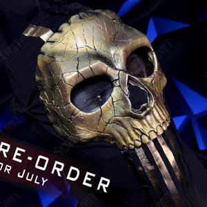 May include: A gold-coloured skull mask with a cracked, weathered texture and deep eye sockets. The mask features vertical gold stripes extending from the jaw area and is attached to a black fabric hood. The text PRE-ORDER FOR JULY is visible.