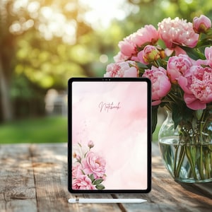 以下が含まれることがあります： ピンク色の水彩デザインに「Notebook」の文字と花のアクセントが施されたデジタルタブレット。ガラスの花瓶に入ったピンクの牡丹のブーケが、木製のテーブルのタブレットの隣に置かれています。白いスタイラスが下に置かれています。