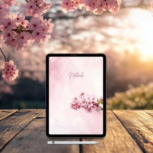 Può includere: Un tablet bianco con uno sfondo acquerellato rosa e bianco. Il testo "Notebook" è scritto in corsivo sullo schermo. Il tablet è appoggiato su un tavolo di legno con una stilo bianca accanto.