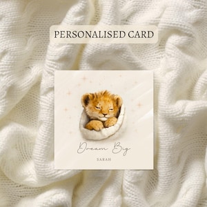May include: A personalised card with a watercolour illustration of a sleeping lion cub in a white blanket. The card reads "Dream Big" and the name "Sarah". The card is set against a white knitted background.