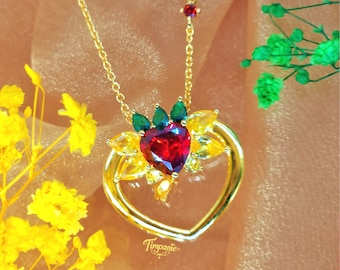 May include: A gold heart-shaped pendant necklace with a red heart-shaped gemstone surrounded by yellow and green gemstones. The necklace is on a gold chain.