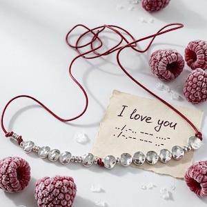 May include: A dark red cord bracelet featuring a series of small, hammered silver-toned circular beads. A rectangular piece of paper with torn edges displays the handwritten text I love you followed by Morse code symbols in black ink.