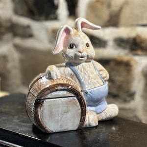 Puede incluir: Una figurita de cerámica de un conejo con un mono azul y sosteniendo un barril de madera. El conejo tiene ojos marrones y un cuerpo marrón claro. La figurita está sentada sobre una superficie de madera oscura.