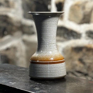 Puede incluir: Un jarrón de cerámica gris con una franja marrón alrededor del centro. El jarrón tiene un cuello estrecho y una base ancha.