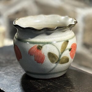 May include: A small, white ceramic pot with a scalloped rim. The pot is decorated with hand-painted red flowers and green leaves.