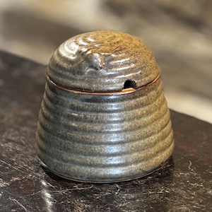 Puede incluir: Un pequeño tarro de miel de cerámica marrón con tapa. El tarro tiene un diseño texturizado en forma de panal.