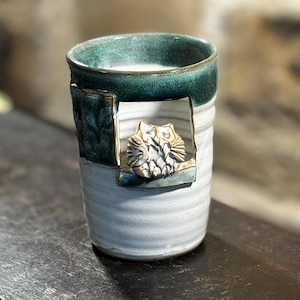 Könnte beinhalten: Eine handgefertigte Keramiktasse mit einem türkisfarbenen Rand und einem strukturierten türkisfarbenen Akzent. Die Tasse zeigt ein Relief von zwei Eulen. Der Körper der Tasse ist hellgrau mit einem weißen Wirbelmuster. Die Tasse steht auf einer dunkelbraunen Oberfläche.