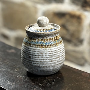 Puede incluir: Un tarro de cerámica blanca con un diseño de rayas marrones y azules y una tapa con pomo blanco. El tarro está sobre una superficie de madera oscura.