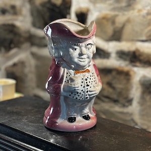 Puede incluir: Un jarro de cerámica con forma de hombre que lleva un sombrero rojo y blanco, una chaqueta roja y una camisa blanca con lunares negros. El jarro tiene una cara blanca con ojos negros y un bigote negro.