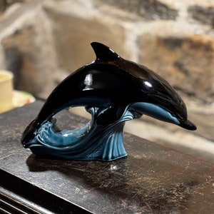 Puede incluir: Una figurita de delfín de cerámica negra y azul saltando fuera del agua. El delfín está en una pose de salto con su cola y aletas extendidas. La figurita está sobre una superficie de madera oscura.