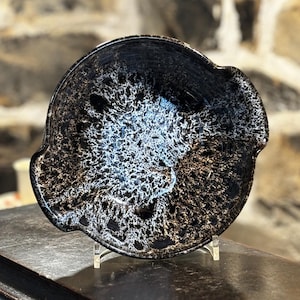 May include: A black ceramic dish with a white and gold speckled glaze. The dish has a rounded shape with two small handles on the sides.