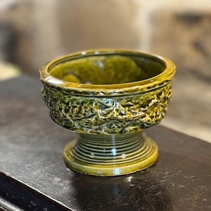 May include: A small, green ceramic planter with a textured, leafy design. The planter has a wide, round base and a narrow, cylindrical top.