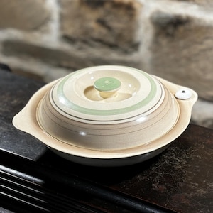 Puede incluir: Una fuente de cerámica con tapa. La fuente es de color beige con rayas verdes y marrones. La tapa tiene un pomo verde. La fuente tiene dos asas y un pequeño detalle blanco en el lateral. La fuente está sobre una superficie oscura.