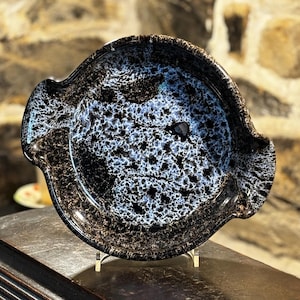 May include: A black and white ceramic serving dish with a speckled glaze. The dish has a rounded shape with two handles on the sides.