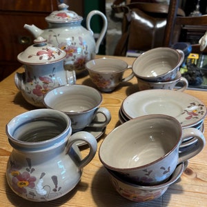 May include: A vintage ceramic tea set with floral designs. The set includes a teapot, creamer, sugar bowl, and saucers. The cups and teapot are a light brown colour with pink and brown floral designs.