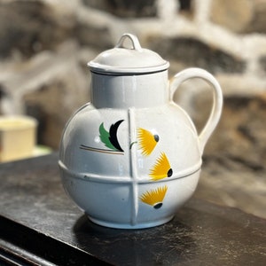 Puede incluir: Jarra de cerámica blanca con un diseño floral amarillo y verde. La jarra tiene un cuerpo redondo y un asa. Está sentada sobre una superficie de madera oscura.