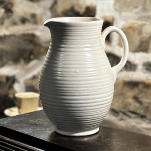 May include: A white ceramic pitcher with a ribbed design and a handle. The pitcher is sitting on a dark wooden surface.