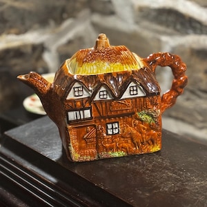 Puede incluir: Una tetera de cerámica marrón y blanca con forma de cabaña con techo de paja. La tetera tiene un pico y una asa y está decorada con ventanas y una puerta. También hay pequeñas figuras de personas y animales en el lateral de la tetera.