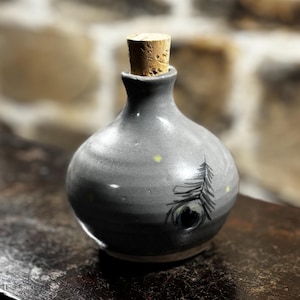 Op de afbeelding: Een klein grijs keramiek flesje met een kurken stop en een geschilderd pauwenveer ontwerp. De fles heeft een gespikkelde grijze glazuur en een witte rand.