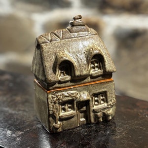 Puede incluir: Una pequeña casa de cerámica con techo de paja, de color marrón y beige, con ventanas y una puerta. La casa está sentada sobre una superficie marrón oscuro.