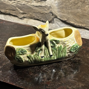 Puede incluir: Un macetero de cerámica con forma de tronco de árbol con dos macetas pequeñas y una figurita de ciervo en la parte superior. El macetero está decorado con hojas verdes y corteza marrón.